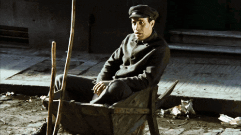a man in a hat sits in a wheelbarrow on the side of the road