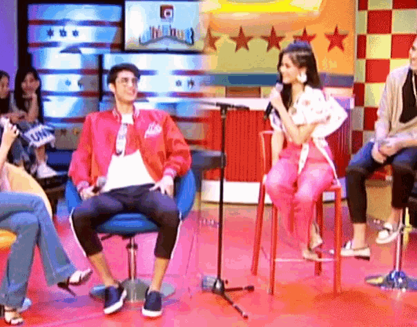 a man in a red jacket sits in front of a microphone while a woman in pink sits on a stool