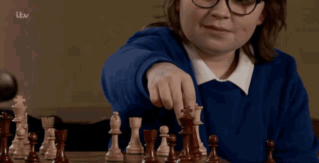 a girl in a blue sweater is playing a game of chess with itv behind her
