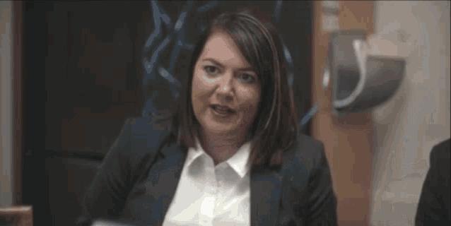 a woman in a suit and white shirt is sitting at a table and says oh my ticking hell .