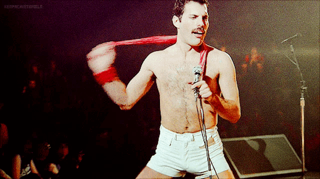 a shirtless freddie mercury is singing into a microphone while holding a red scarf around his neck
