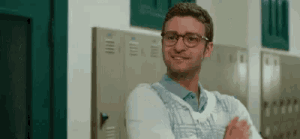 a man wearing glasses and a sweater is standing in front of lockers in a hallway .
