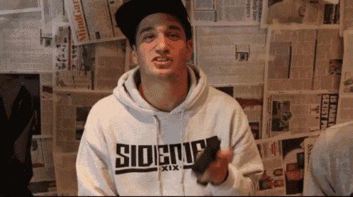 a man wearing a sideup hoodie stands in front of a wall covered in newspapers