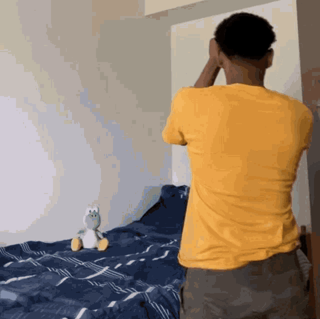 a man in a yellow shirt is standing in front of a stuffed animal on a bed