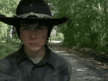 a young man wearing a cowboy hat stands on a dirt road in the woods