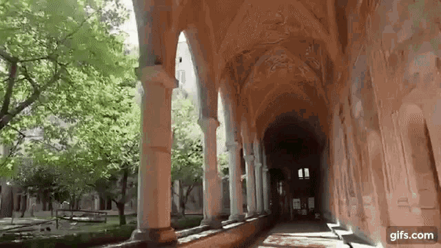 a long hallway with columns and arches leading to a building surrounded by trees .