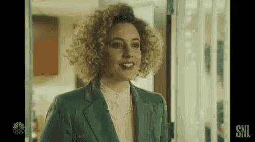 a woman with curly hair is standing in a doorway wearing a green suit and a pink shirt .