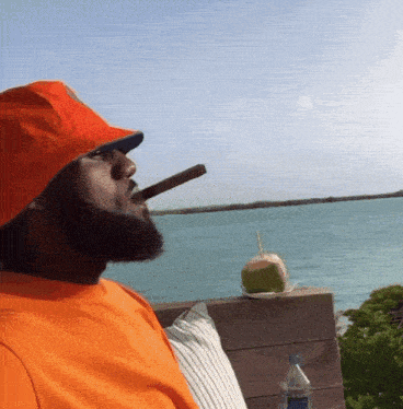 a man in an orange shirt is smoking a cigar in front of a body of water