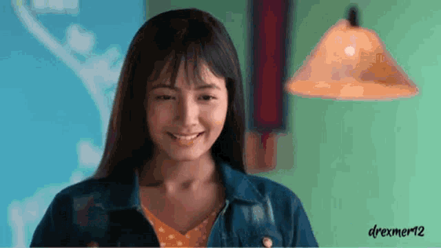 a young woman in a denim jacket is smiling in a room with a lamp in the background .