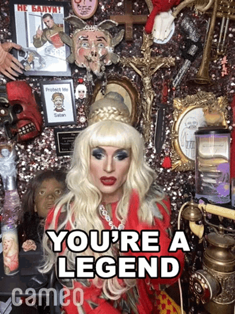 a drag queen says " you 're a legend " in front of a wall of memorabilia