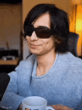 a man wearing sunglasses and a blue sweater is holding a cup of coffee and smiling .