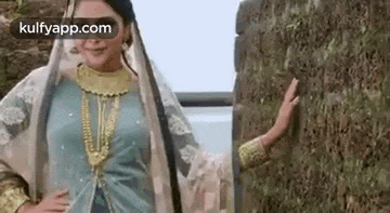 a woman in a blue dress and gold jewelry is standing next to a mossy wall .