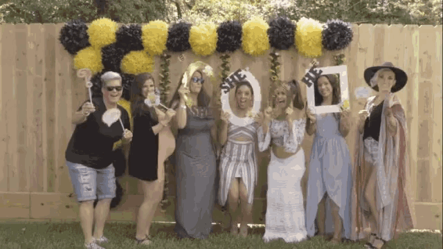 a group of women standing in front of a fence holding props including one that says she be