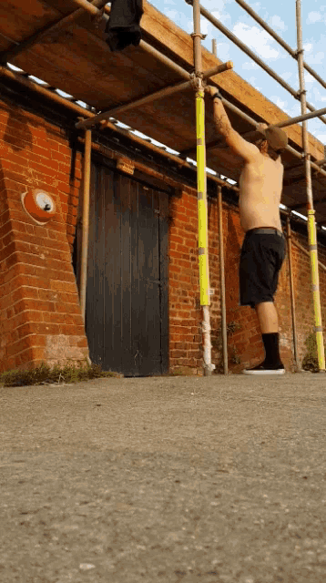 a shirtless man stands on a scaffolding in front of a brick wall