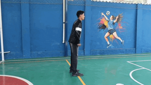 a man is standing on a basketball court with a mural of a volleyball player on the wall behind him .