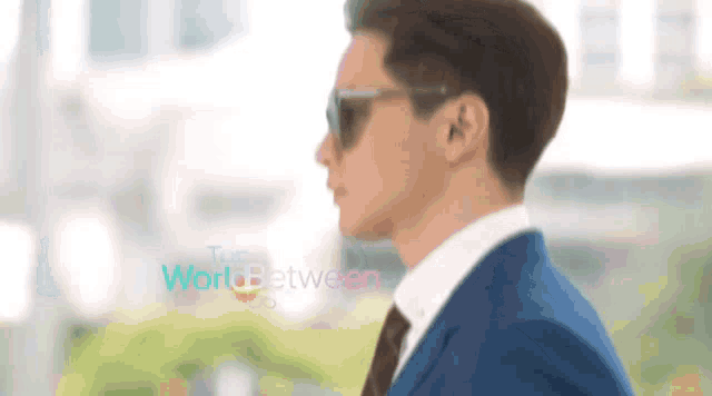 a man wearing sunglasses and a suit is standing in front of a sign that says the world between
