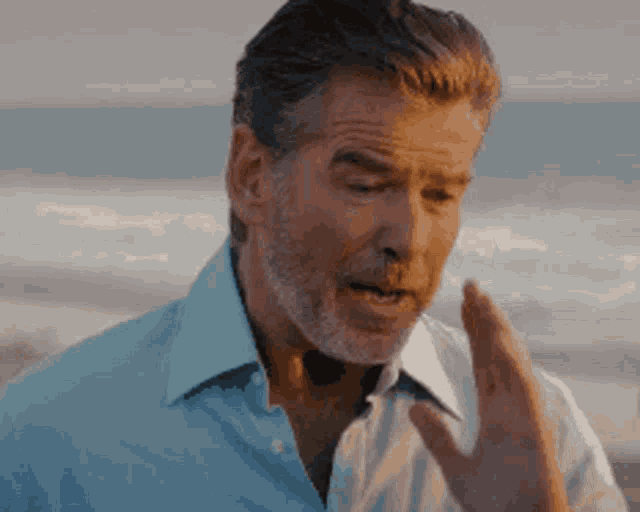 a man with a beard and a blue shirt is standing on a beach talking on a cell phone .