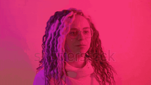 a woman with dreadlocks and glasses is standing in front of a pink background and looking at the camera .