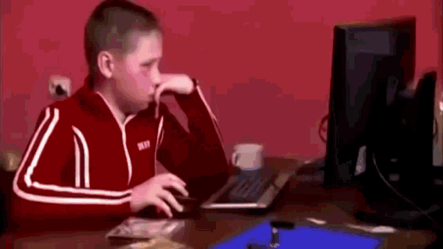 a young boy in a red jacket is sitting at a desk in front of a computer monitor .