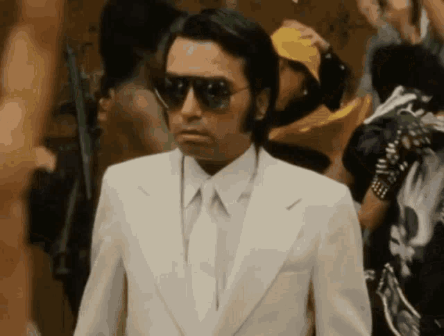 a man wearing sunglasses and a white suit stands in a crowd