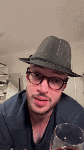 a man wearing glasses and a fedora holds a glass of wine