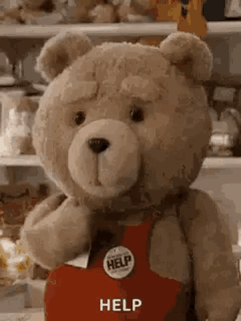 a teddy bear wearing a red apron with a help button on it is standing in front of a refrigerator .