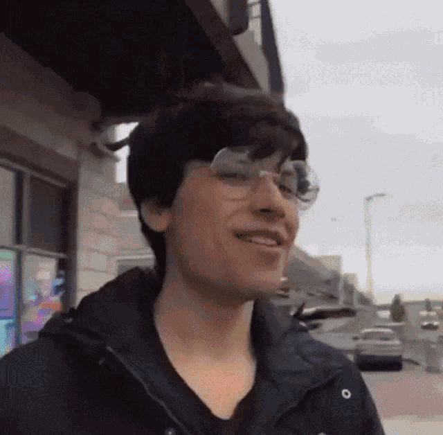 a young man wearing glasses and a black jacket is smiling and looking at the camera .