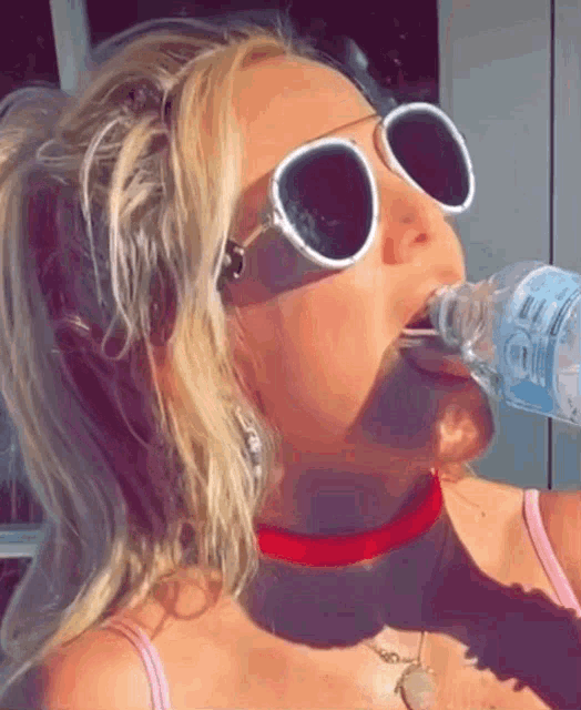 a woman wearing sunglasses and a red choker drinks from a bottle of water