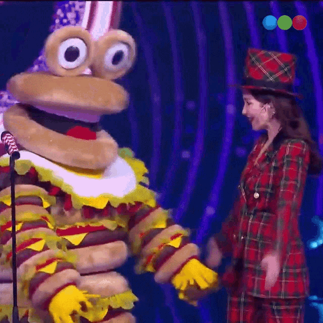 a woman in a plaid suit stands next to a hamburger mascot in front of a microphone