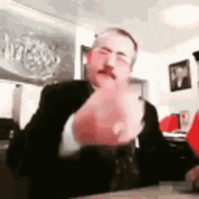 a man with a mustache and glasses is sitting at a table with his hand up .