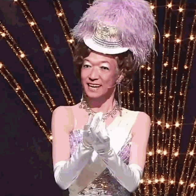 a woman wearing a feathered hat and gloves is clapping her hands on a stage .