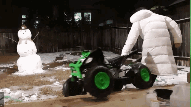 a person in a white coat is pushing a green and black atv in front of a snowman
