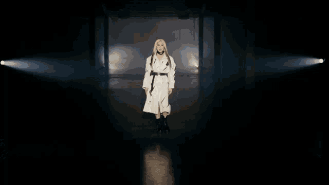 a woman in a white trench coat and black boots stands in a dark room