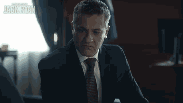 a man in a suit and tie is sitting in front of a sign that says " jack ryan "