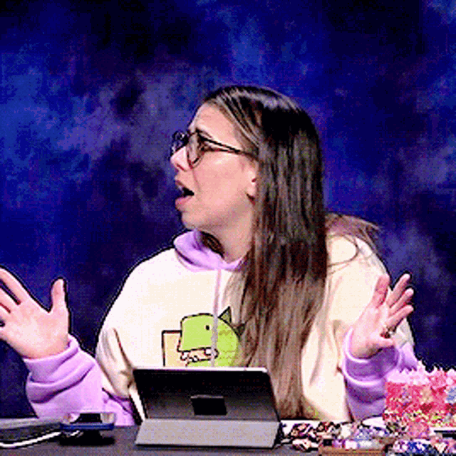 a woman wearing glasses and a hoodie with a dinosaur on it is sitting at a table with a tablet