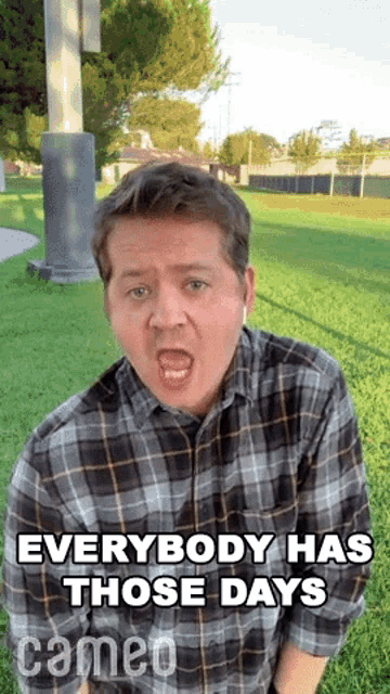 a man in a plaid shirt is making a funny face in a park while wearing ear buds .