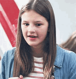 a young girl wearing a striped shirt and a denim jacket is looking at the camera .