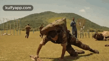 a soldier is doing push ups on the ground in a field while other soldiers run in the background .