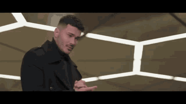 a man in a black coat and black turtleneck is standing in front of a wall with lights .