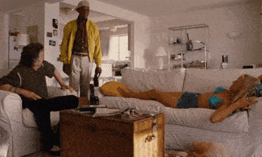 a man in a yellow jacket is standing next to a woman laying on a white couch