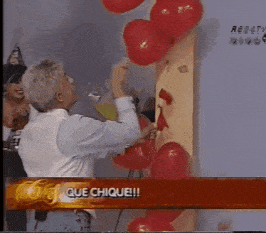 a man in a white shirt is holding a red balloon in front of a sign that says que chique