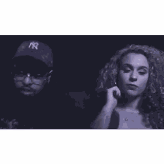 a man and a woman are standing next to each other in a dark room . the man is wearing a ny hat .