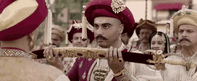 a man with a mustache is holding a sword in front of a crowd of people wearing turbans .