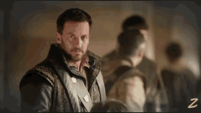 a man with a beard is standing in front of a group of people with the letter z in the corner