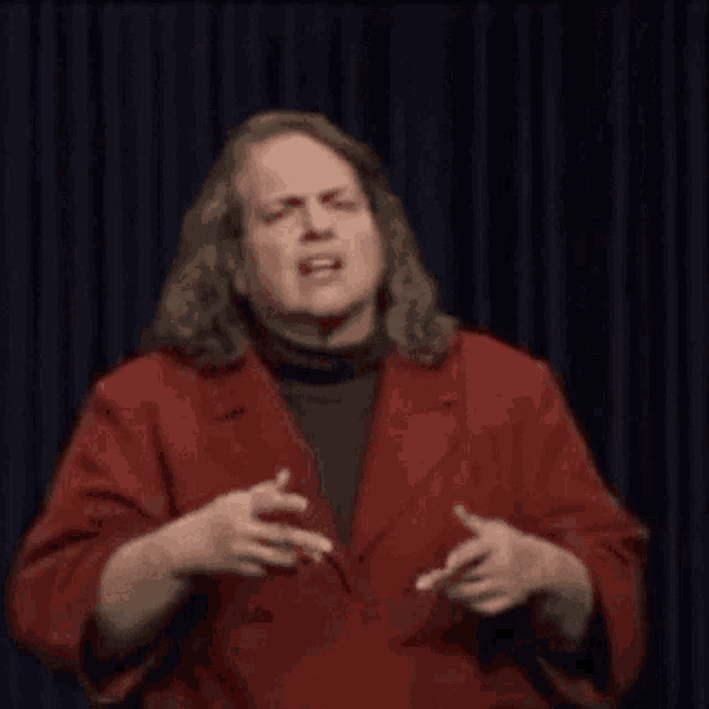 a man with long hair is wearing a red jacket and making a funny face while talking on a stage .