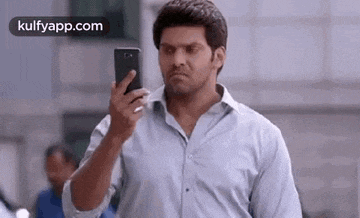 a man in a blue shirt is holding a cell phone in his hand and looking at it .