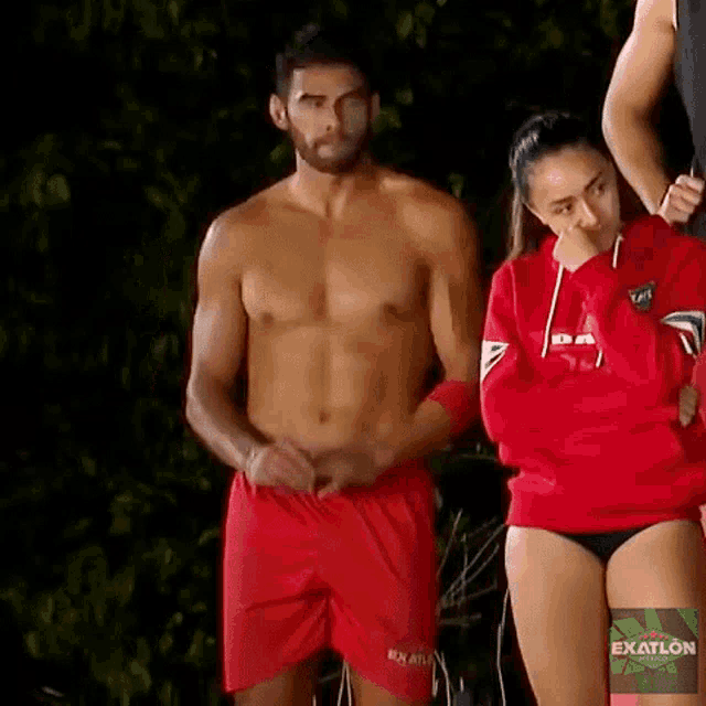 a shirtless man in red shorts is standing next to a woman in a red sweatshirt with the word exatlon on it