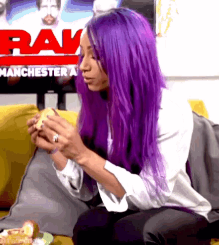 a woman with purple hair sits on a couch eating a sandwich