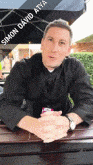 a man sits at a table under an umbrella with the name simon david atya on his shirt