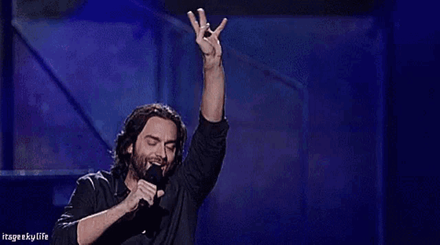 a man stands on a stage holding a microphone with his hand in the air and the words itsgeekylife above him